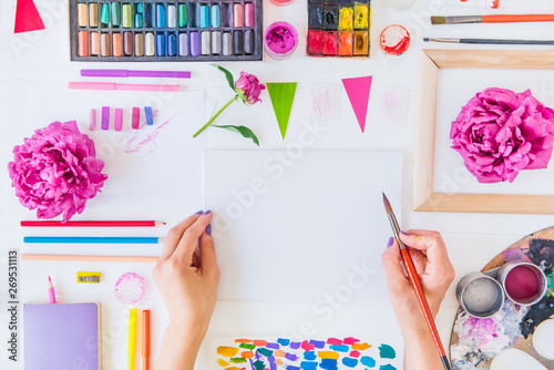 Top view Female Hands holding painting brush over blank canvas with paintiing materials and flowers on white wooden background. Add more colors to your life, be cheerful motivated inspired Concept. photo