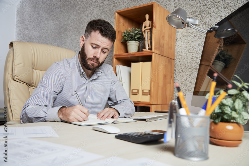 Bearded young manager sitting at office desk and writing new plans and ideas in his notepad Bearded young manager sitting at office desk and writing new plans and ideas in his notepad