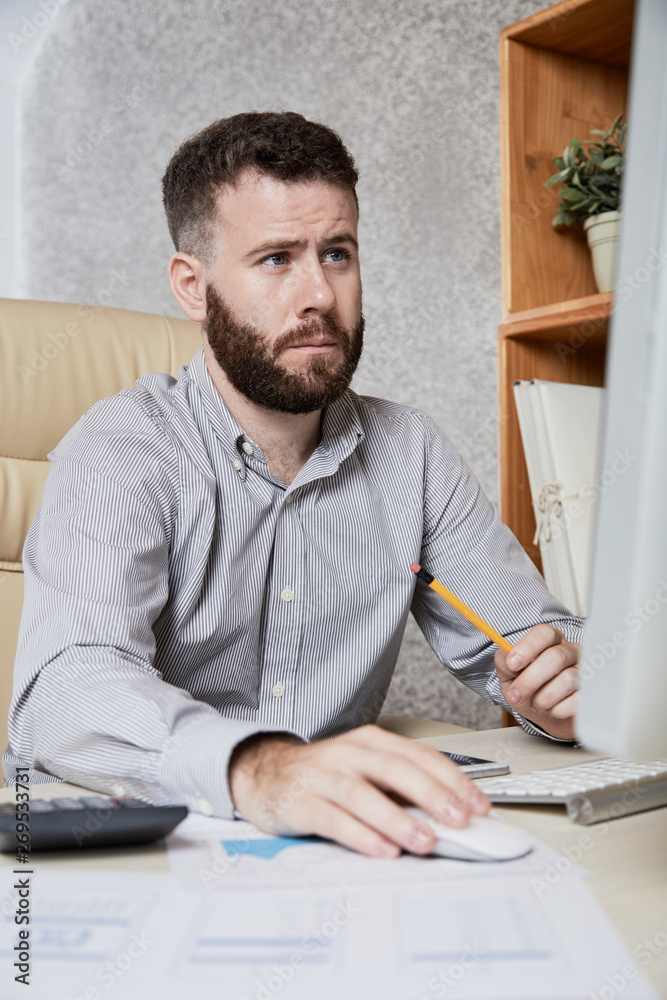 Bearded serious manager concentrating on his work on computer while sitting at workplace at office