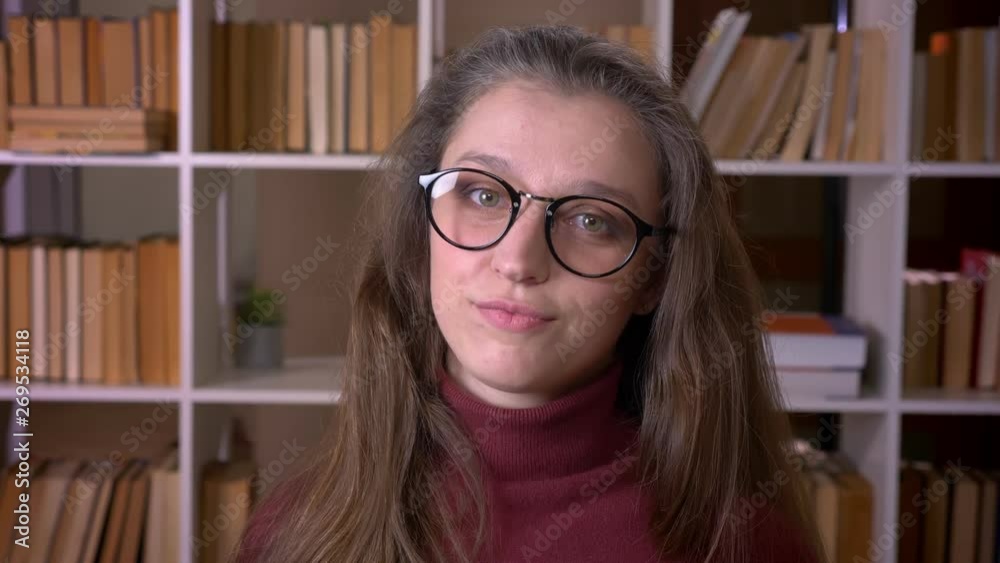 Closeup portrait of young caucasian female student in glasses nodding saying yes smiling happily looking at camera in the college library indoors Closeup portrait of young caucasian female student in glasses nodding saying yes smiling happily looking at camera in the college library indoors