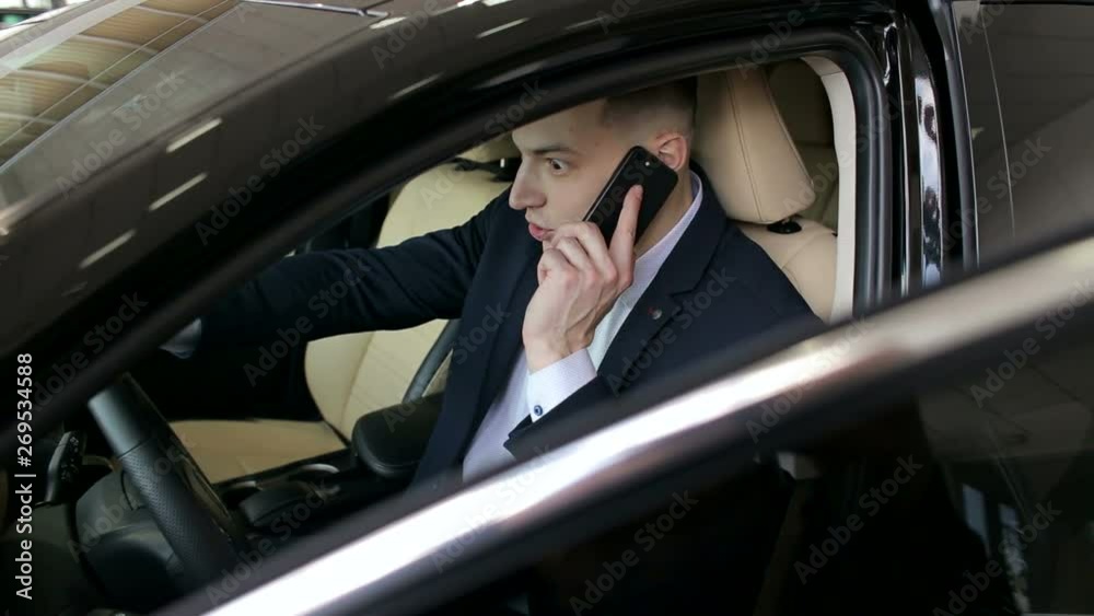 A successful young businessman in a suit talking on the phone while sitting behind the wheel in his new car in the showroom. Portrait of a businessman in a car with a phone. A successful young businessman in a suit talking on the phone while sitting behind the wheel in his new car in the showroom. Portrait of a businessman in a car with a phone.