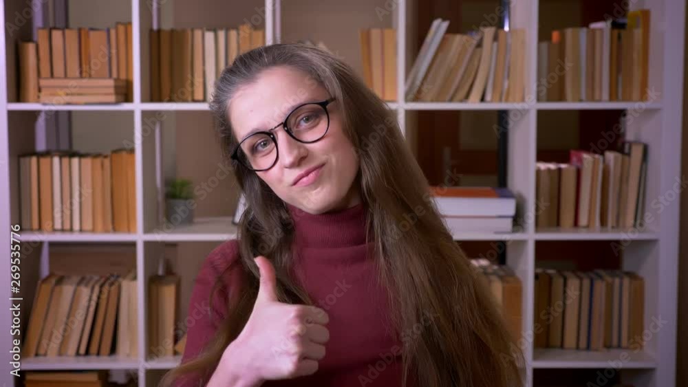 Closeup portrait of young caucasian female student in glasses showing a thumb up smiling happily looking at camera in the college library indoors