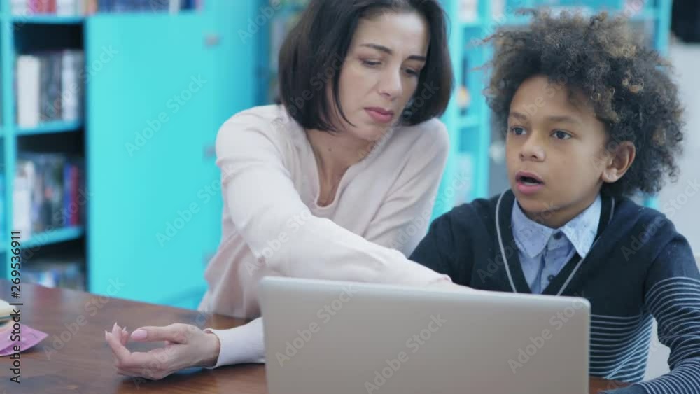 Caucasian female teacher explaining to mixed race boy how to solve task on laptop computer sitting at desk in classroom, tracking right medium shot
