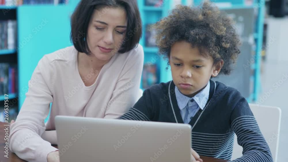 Middle aged female teacher explaining to upset mixed race school boy how to work on laptop computer sitting at desk in classroom, tracking right medium shot Middle aged female teacher explaining to upset mixed race school boy how to work on laptop computer sitting at desk in classroom, tracking right medium shot