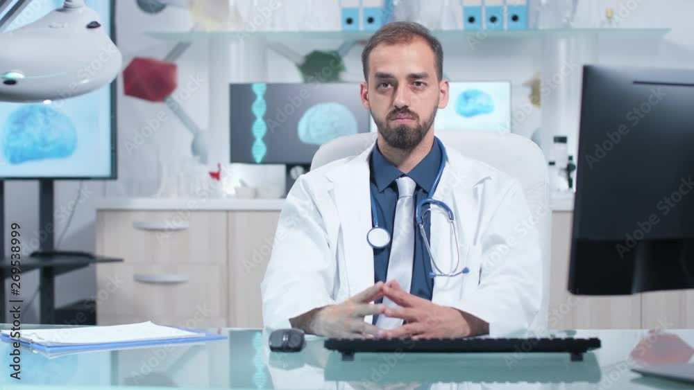Portrait of serious doctor looking at the camera while sitting in his desk in modern office or laboratory. There are 3D animation of a brain running in the background on the monitors Portrait of serious doctor looking at the camera while sitting in his desk in modern office or laboratory. There are 3D animation of a brain running in the background on the monitors