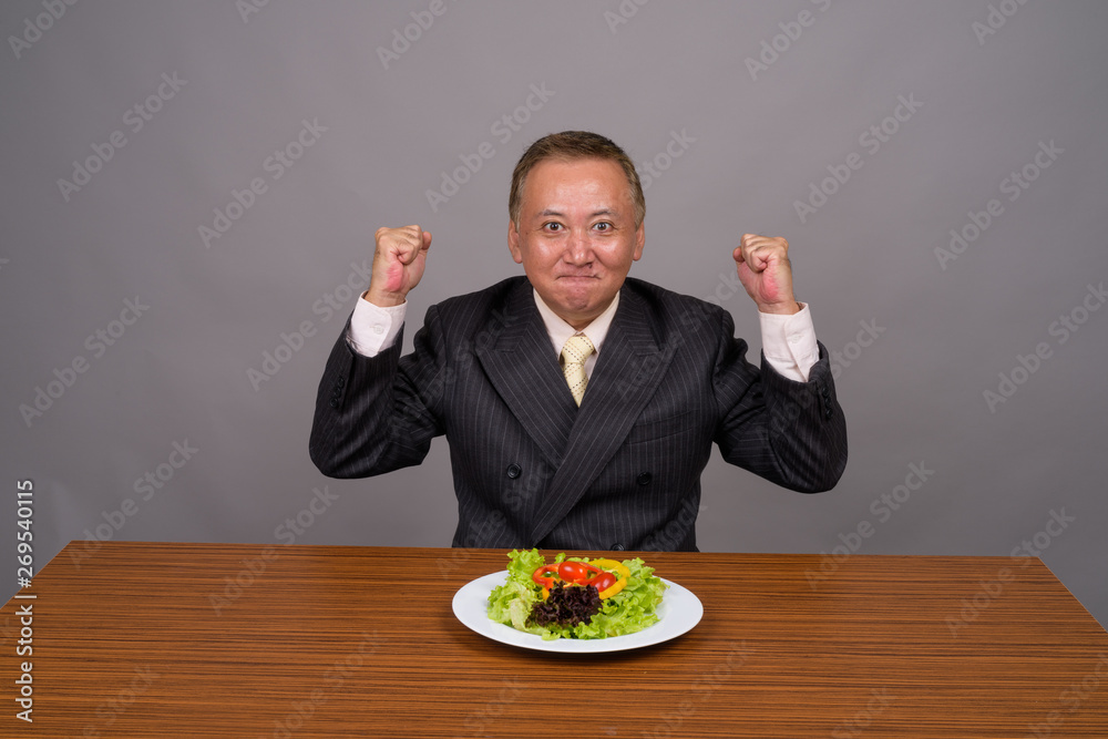 Mature Asian businessman sitting with wooden table against gray Mature Asian businessman sitting with wooden table against gray