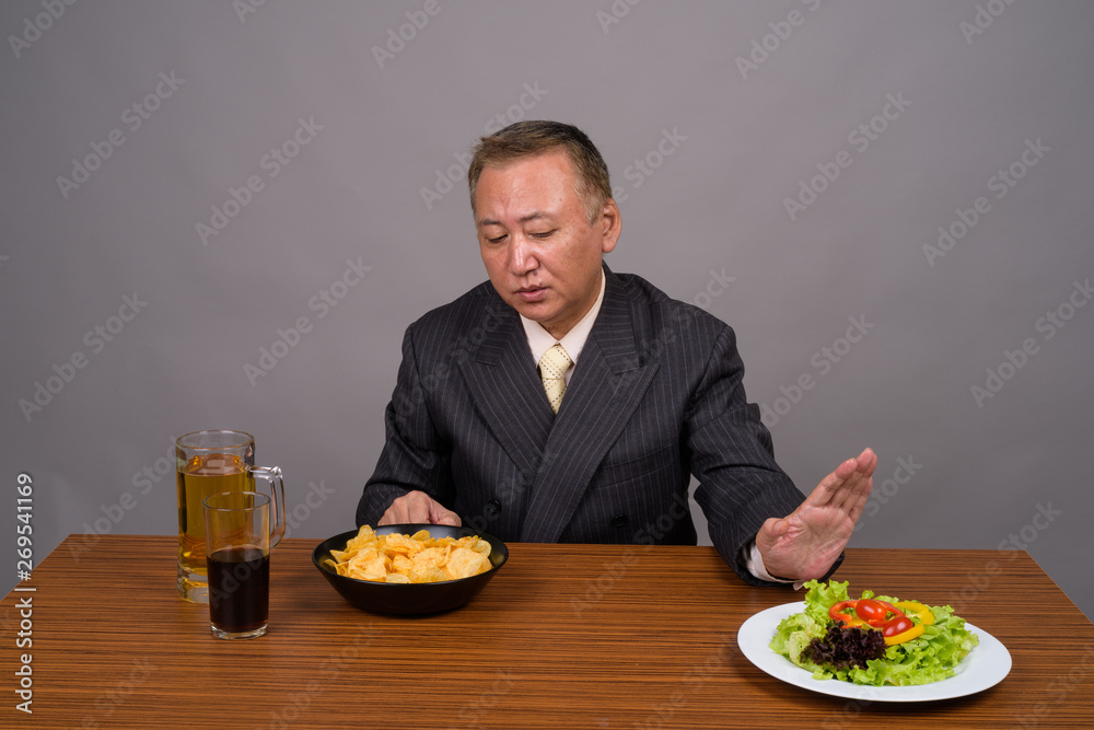Mature Asian businessman sitting with wooden table against gray Mature Asian businessman sitting with wooden table against gray