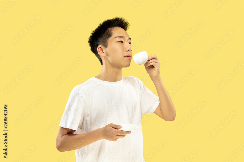 Korean young man's half-length portrait on yellow studio background. Male model in white shirt. Drinking coffee, feeling happy. Concept of human emotions, facial expression. Front view. Trendy colors. Korean young man's half-length portrait on yellow studio background. Male model in white shirt. Drinking coffee, feeling happy. Concept of human emotions, facial expression. Front view. Trendy colors.