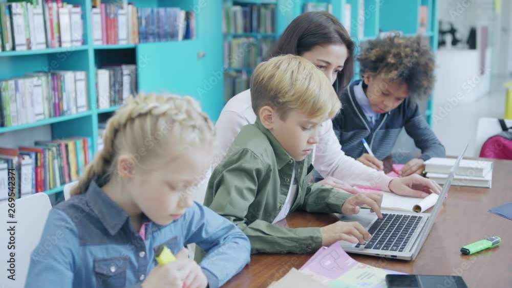 Tracking right shot of female teacher helping schoolboy to study on laptop computer. Two elementary students, boy and girl, sitting at desk with them and writing on sticky notes Tracking right shot of female teacher helping schoolboy to study on laptop computer. Two elementary students, boy and girl, sitting at desk with them and writing on sticky notes