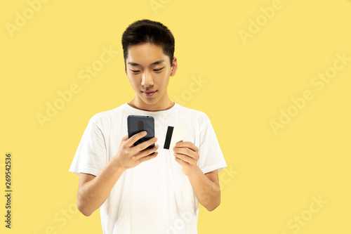 Korean young man's half-length portrait on yellow studio background. Male model in white shirt. Paying bill or buying online. Concept of human emotions, facial expression. Front view. Trendy colors. © master1305