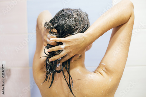 Young happy woman washing her hair with shampoo, foaming with hands. Beautiful brunette girl taking shower and enjoying relax time. Body, hair and skin hygiene, lifestyle concept.