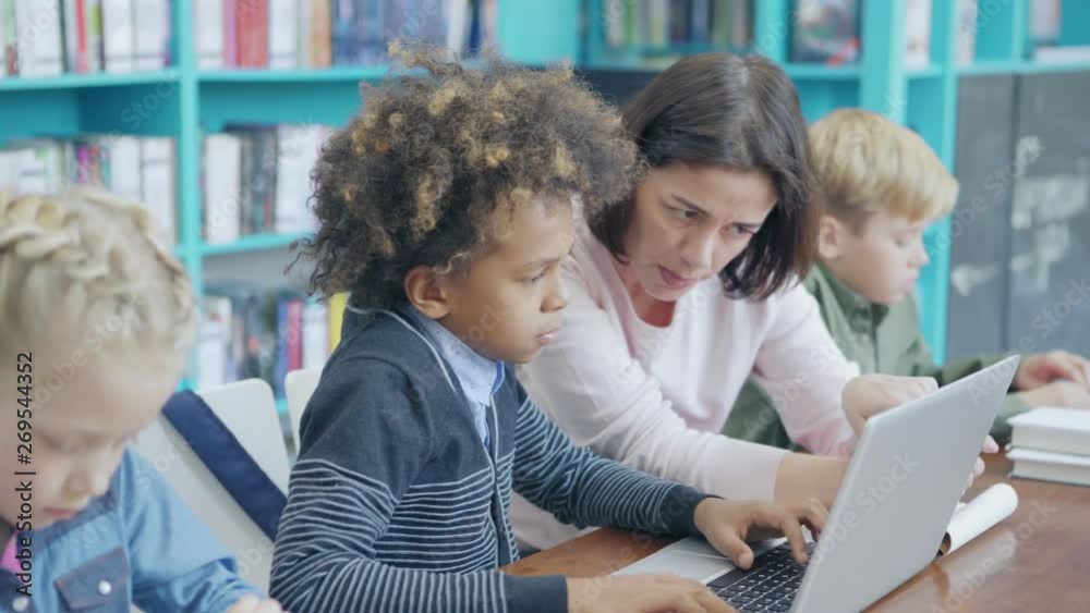 Tracking right shot of middle aged female teacher and group of diverse elementary students sitting at desk in classroom. Tutor explaining mixed race schoolboy how to work on laptop computer