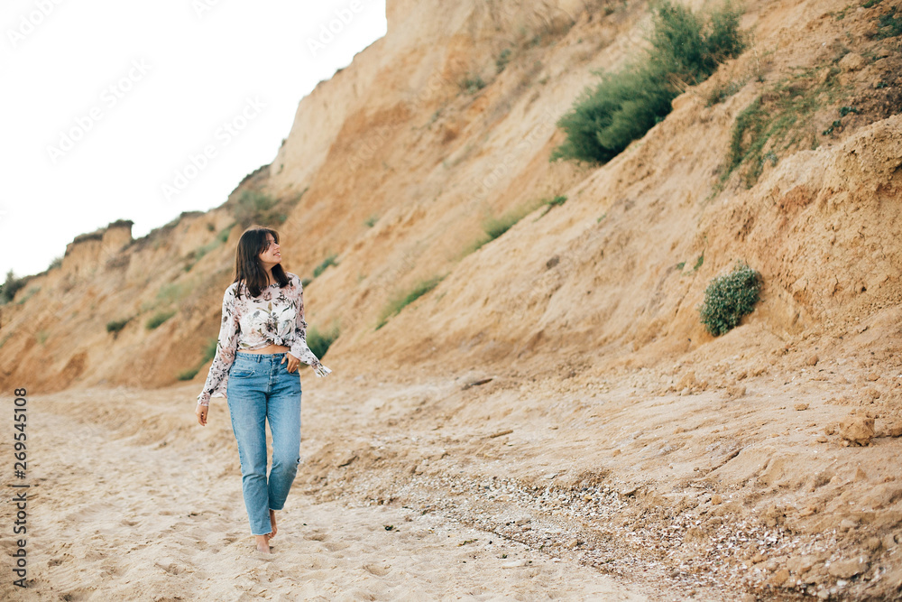 Naklejka premium Stylish hipster girl walking on beach at sea and relaxing. Happy boho woman in denim jeans and floral blouse relaxing at sandy cliff on tropical island. Travel and summer vacation concept