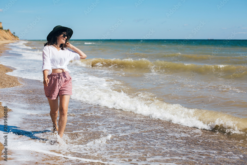 Summer vacation. Happy young boho woman walking in sea waves in sunny warm day at tropical island and blue sky. Space for text. Stylish hipster girl in hat relaxing on beach and smiling. Summer vacation. Happy young boho woman walking in sea waves in sunny warm day at tropical island and blue sky. Space for text. Stylish hipster girl in hat relaxing on beach and smiling.