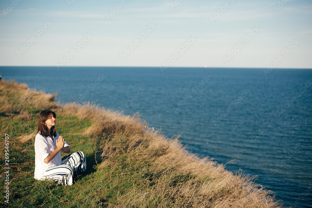 Yoga day. Stylish hipster girl sitting in yoga pose and relaxing on mountain with sea view. Happy boho woman practicing yoga and meditation on tropical island. Mindful concept. Space for text