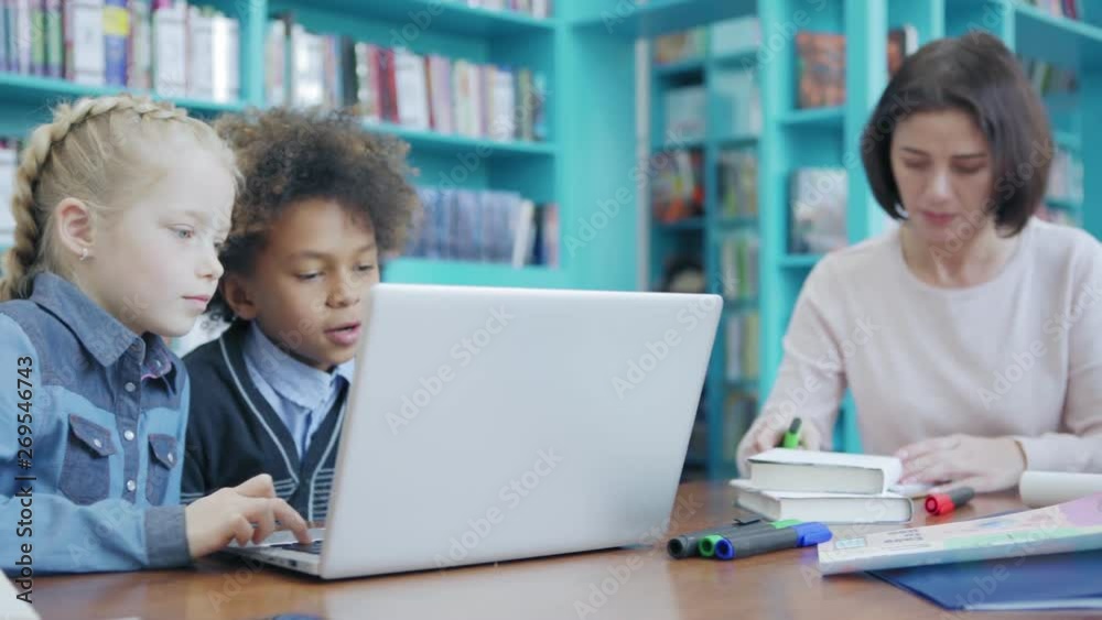 Teacher helping elementary school boy to do homework while two students having fun playing on laptop computer sitting at desk nearby, tracking shot Teacher helping elementary school boy to do homework while two students having fun playing on laptop computer sitting at desk nearby, tracking shot