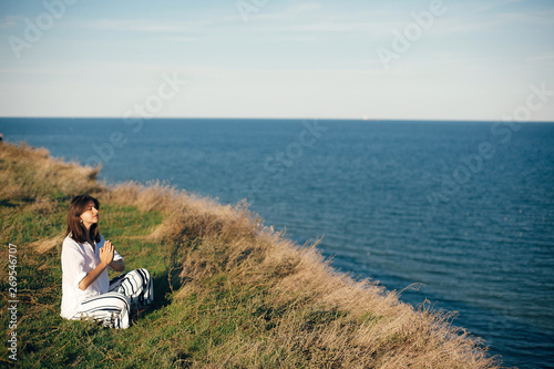 Yoga day. Stylish hipster girl sitting in yoga pose and relaxing on mountain with sea view. Happy boho woman practicing yoga and meditation on tropical island. Mindful concept. Space for text