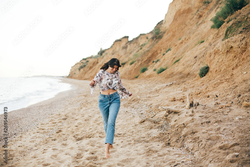 Stylish hipster girl running on beach at sea and smiling. Happy fashionable boho woman relaxing at sandy cliff on tropical island. Travel and summer vacation. Space for text
