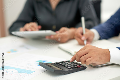 Close-up of businessman sitting at office desk with financial graphs and calculating incomes of the company with businesswoman using tablet pc near by him