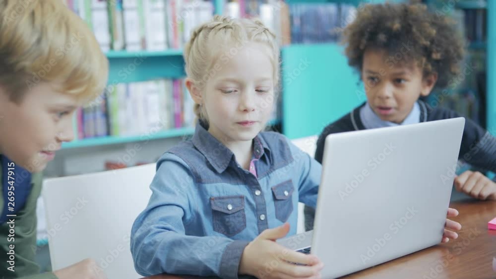 Cute schoolgirl sitting at desk and doing task on laptop computer. Two boys joining her in elementary school classroom to help and advise Cute schoolgirl sitting at desk and doing task on laptop computer. Two boys joining her in elementary school classroom to help and advise