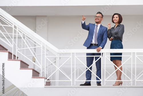 Smiling Asian businessman in suit pointing at something while standing together with young businesswoman on staircase of modern office