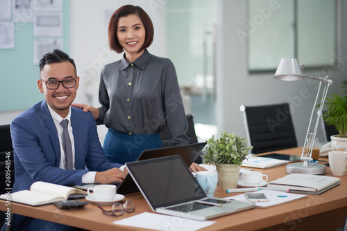 Portrait of Asian young businessman sitting at office desk typing on laptop with beautiful businesswoman standing near by him, they smiling at camera at office