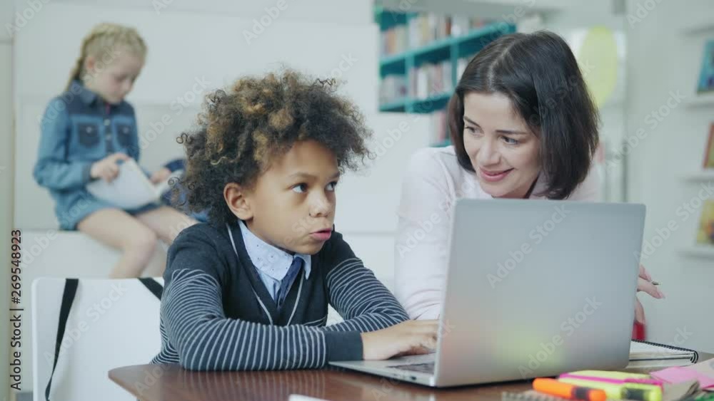 Rack focus of blonde schoolgirl reading book while teacher explaining task on laptop computer to his male classmate sitting at desk in elementary school classroom Rack focus of blonde schoolgirl reading book while teacher explaining task on laptop computer to his male classmate sitting at desk in elementary school classroom