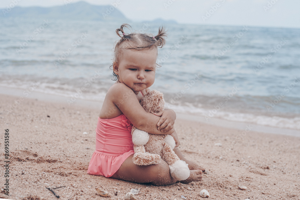 pensive little girl hugging teddy bear and looking away while sitting on seashore. pensive little girl hugging teddy bear and looking away while sitting on seashore.