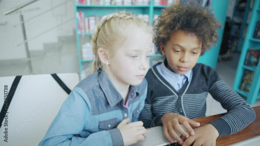 Two elementary school kids, blonde girl and mixed race boy, having fun and laughing while playing on laptop computer sitting at desk together Two elementary school kids, blonde girl and mixed race boy, having fun and laughing while playing on laptop computer sitting at desk together