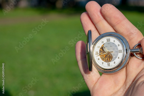 Vintage pocket watch in male hand on a background of green grass. Steampunk watch. Sunny summer day. The clock mechanism is partially visible