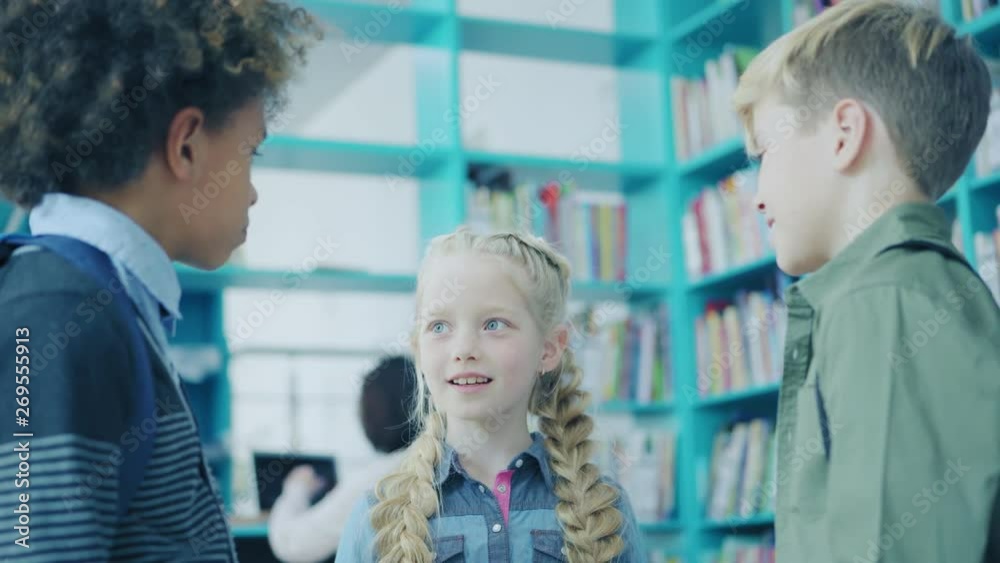 Group of three elementary students standing together and talking near bookshelves in school library. Focus on beautiful schoolgirl with blonde braids, tracking right medium shot
