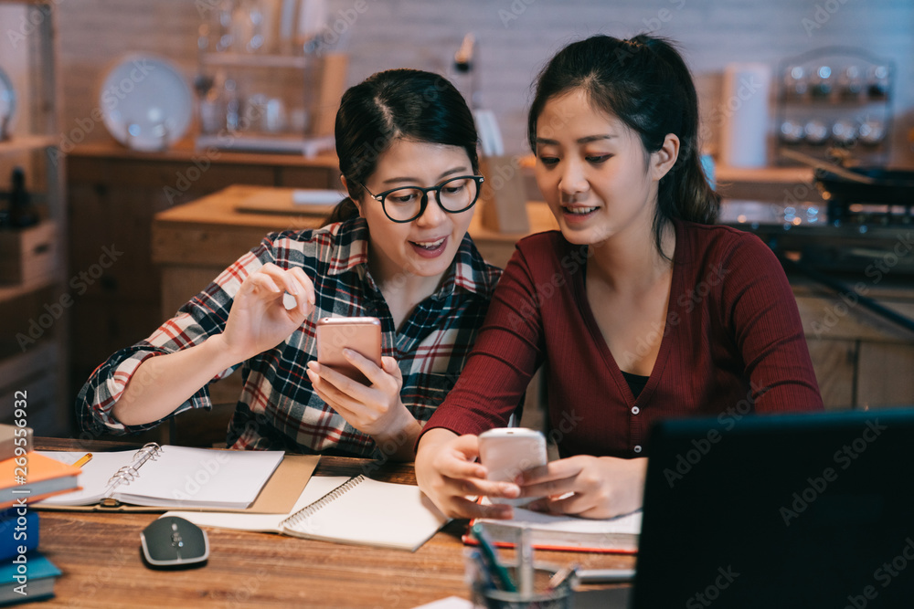 Φωτογραφία two college asian girls friends sitting at wooden home kitchen table in midnight using cellphone relaxing