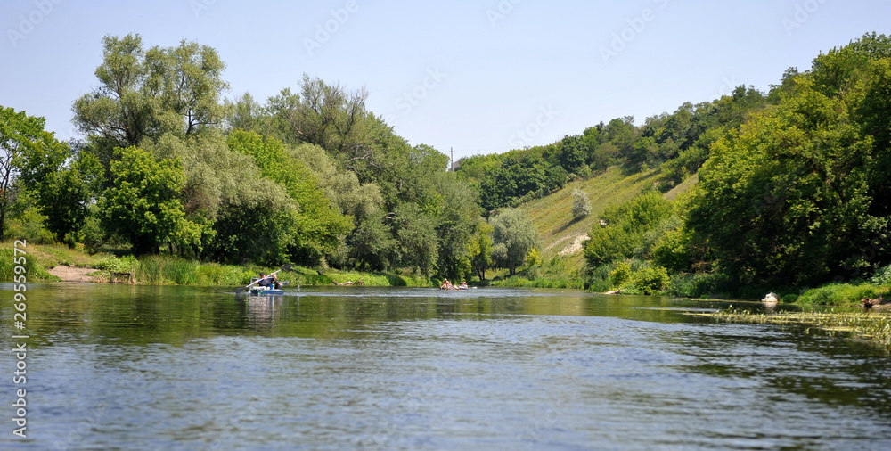 A picture of the tourists on a sunny summer day canoeing on the river. Green trees grow on the banks of the river. Panorama of the boat with oarsmen on the river.