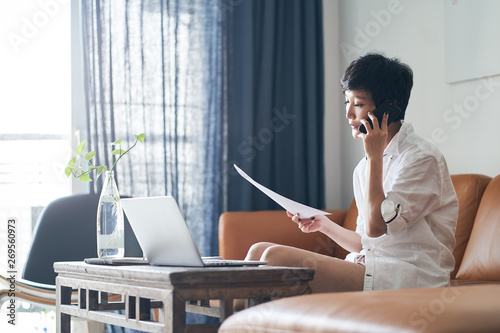 Photography Asian self employed woman sitting on couch  talking on phone & working on laptop