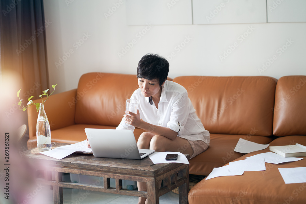 Asian self employed woman sitting on couch  & working on laptop at home