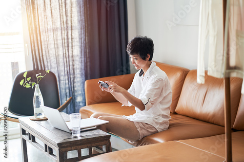 Asian self employed woman sitting on couch taking photo with smartphone while working on laptop at home