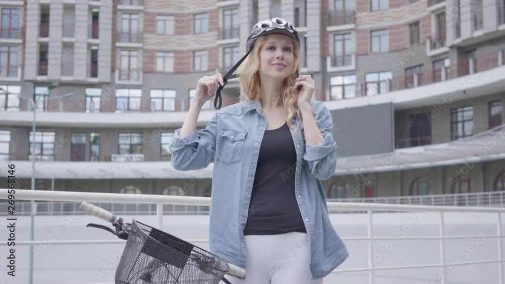 Pretty blond woman putting a bike helmet on her head and riding her bicycle against the background of urban architecture. The lady on cycle track in the city in front of skyscraper. Active lifestyle Pretty blond woman putting a bike helmet on her head and riding her bicycle against the background of urban architecture. The lady on cycle track in the city in front of skyscraper. Active lifestyle