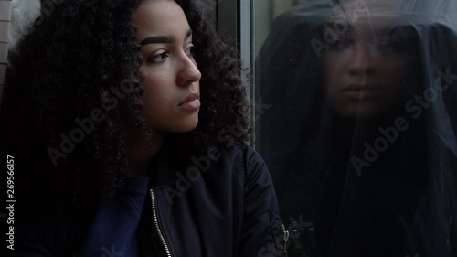 Sad depressed beautiful female mixed race African American girl teenager young woman teen wearing a black jacket sitting looking out of a window 