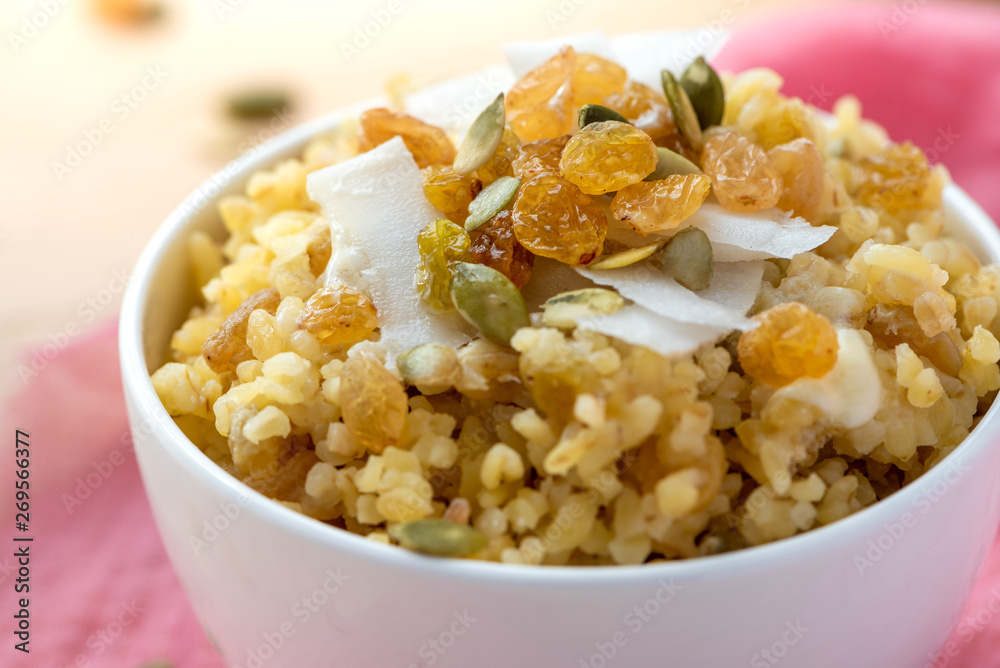 Cooked bulgur with raisins, seeds and coconut in a white bowl on a wooden table. Sweet bulgur porridge close up, Turkish cuisine. Cooked bulgur with raisins, seeds and coconut in a white bowl on a wooden table. Sweet bulgur porridge close up, Turkish cuisine.