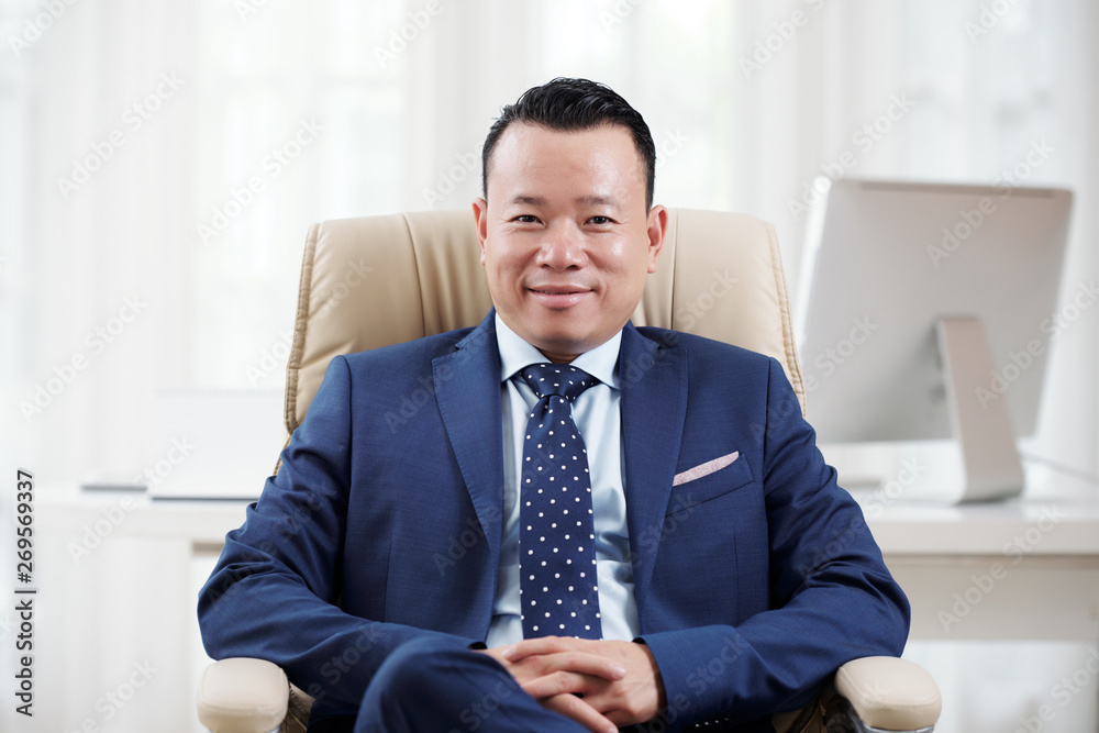 Portrait of Asian mature businessman sitting on chair in blue suit looking at camera and smile while working at office Portrait of Asian mature businessman sitting on chair in blue suit looking at camera and smile while working at office