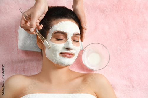 Cosmetologist applying white mask onto woman's face in spa salon, top view