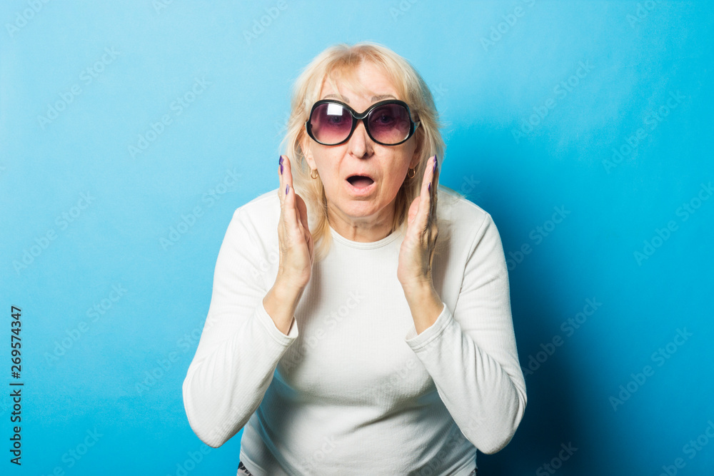 Old woman wearing sunglasses with a surprised face makes a gesture with her hands against a blue background. Concept of surprise, shock, unexpected news Old woman wearing sunglasses with a surprised face makes a gesture with her hands against a blue background. Concept of surprise, shock, unexpected news