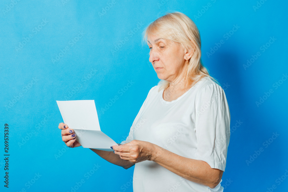 Old woman is holding in her hands a clean white sheet of paper on a blue background. The concept of receiving letters, notifications, invoices for payment, taxes Old woman is holding in her hands a clean white sheet of paper on a blue background. The concept of receiving letters, notifications, invoices for payment, taxes