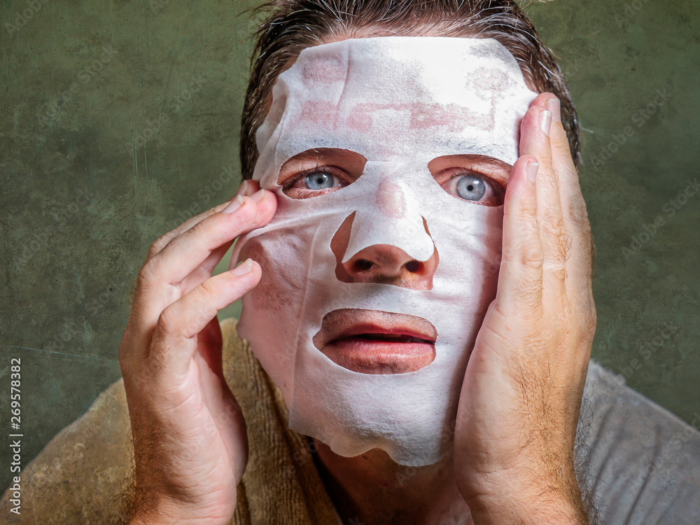Fototapeta premium young funny and messy man applying beauty facial mask looking to the mirror surprised and horrified in skin care aging concept and male cosmetic face treatment