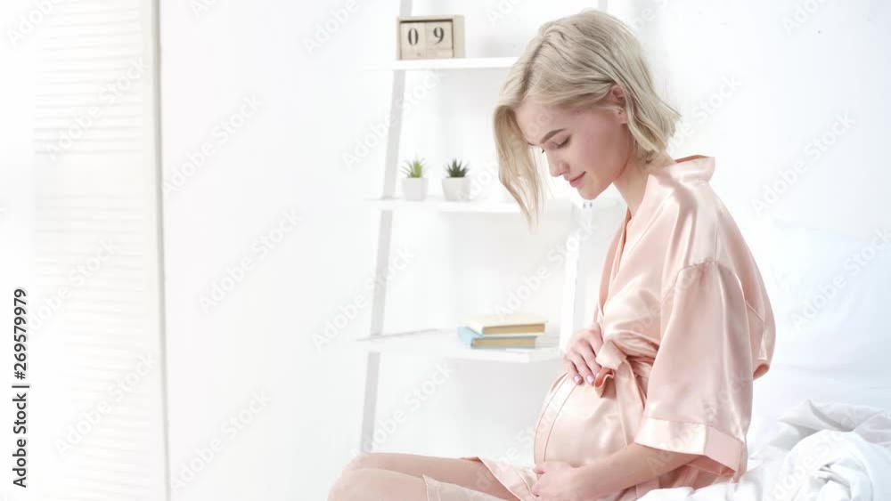cheerful pregnant woman touching belly and smiling while sitting in bedroom cheerful pregnant woman touching belly and smiling while sitting in bedroom