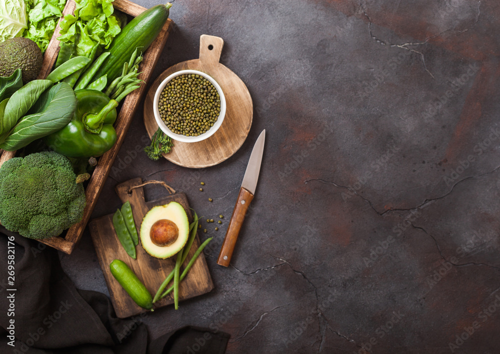 Assorted green toned raw organic vegetables in wooden box on dark background. Avocado, cabbage, cauliflower and cucumber with trimmed and mung beans and chopping board with knife. Space for text Assorted green toned raw organic vegetables in wooden box on dark background. Avocado, cabbage, cauliflower and cucumber with trimmed and mung beans and chopping board with knife. Space for text