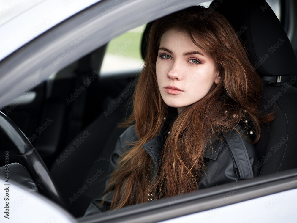 Close up portrait of young attractive red hair self-employed business woman driver sitting in white car stuck in a city traffic jam staring into camera running late to work noonday bleached colors