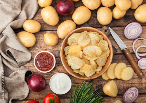 Fresh organic homemade potato crisps chips in wooden bowl with sour cream and red onions and spices on wooden table background. Top view.Fresh yellow potatoes with ketchup