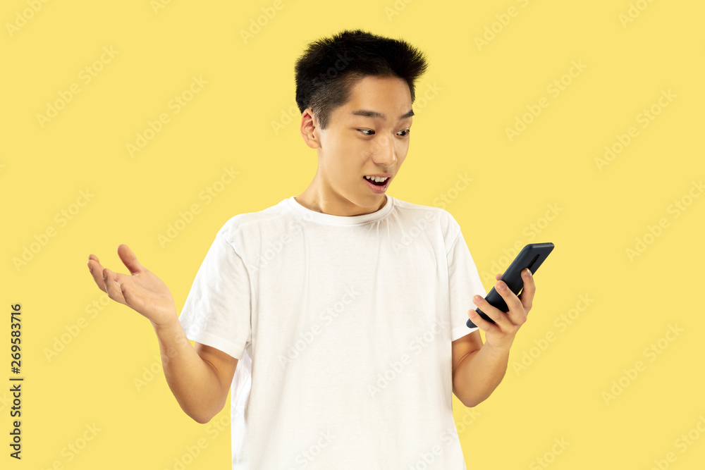 Korean young man's half-length portrait on yellow studio background. Male model in white shirt. Using smartphone for betting, news reading or talking. Concept of human emotions, facial expression. Korean young man's half-length portrait on yellow studio background. Male model in white shirt. Using smartphone for betting, news reading or talking. Concept of human emotions, facial expression.