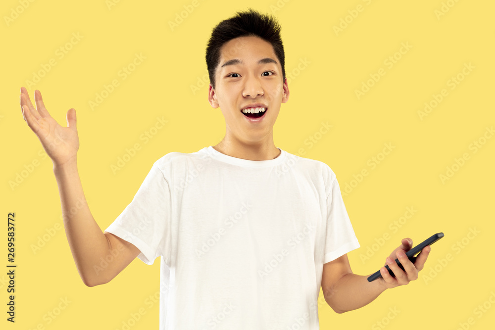 Korean young man's half-length portrait on yellow studio background. Male model in white shirt. Using smartphone for betting, news reading or talking. Concept of human emotions, facial expression. Korean young man's half-length portrait on yellow studio background. Male model in white shirt. Using smartphone for betting, news reading or talking. Concept of human emotions, facial expression.